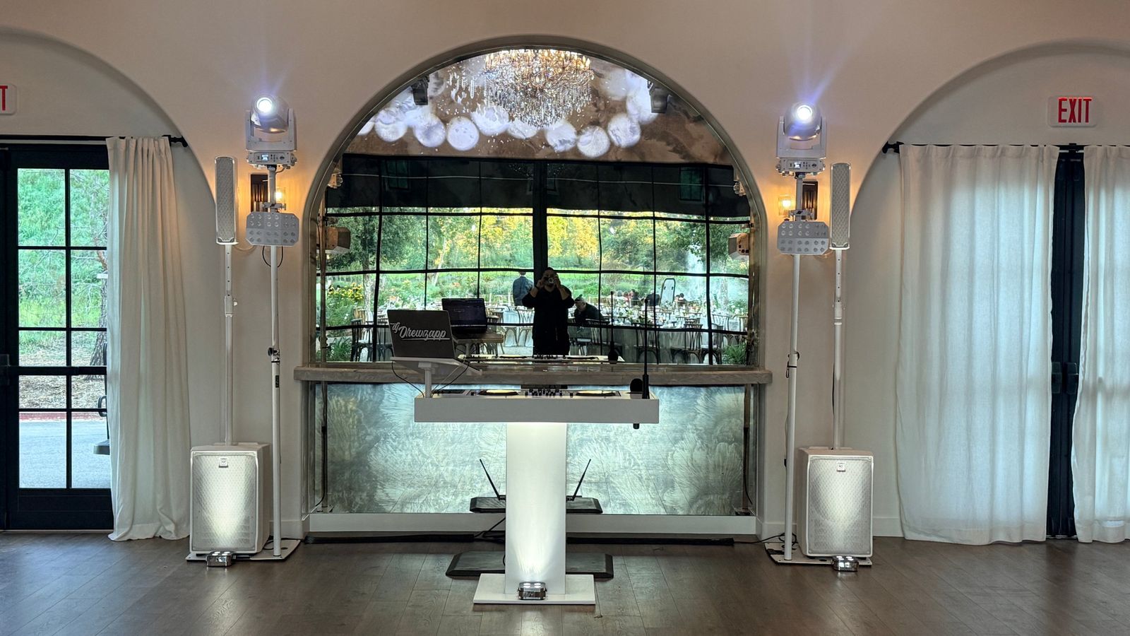 White DJ booth set up in front of an arched mirror, flanked by white column speakers and moving-head lights