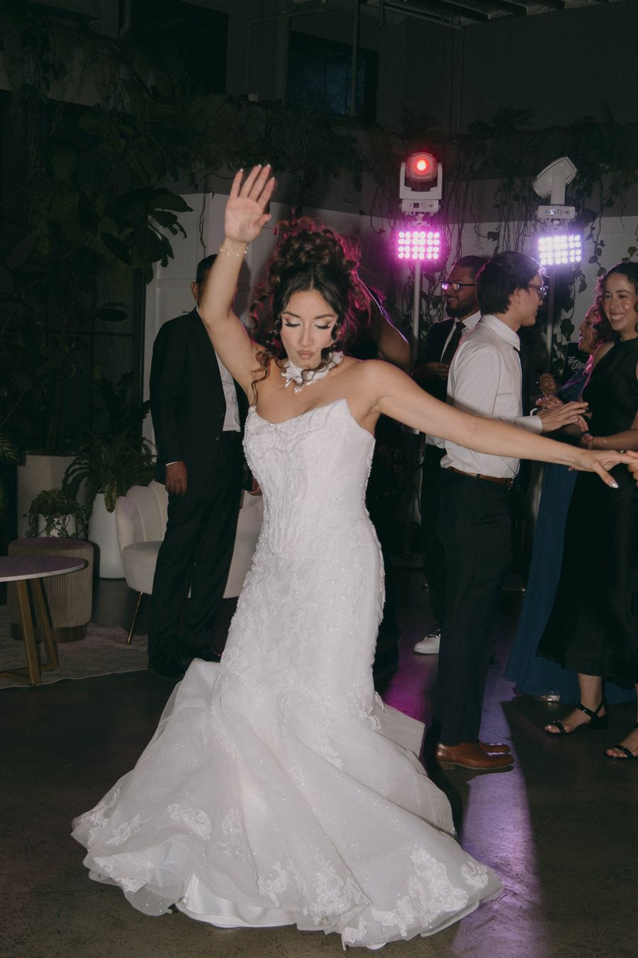 A bride spins on the dance floor with one arm raised, guests dancing behind her under pink lights